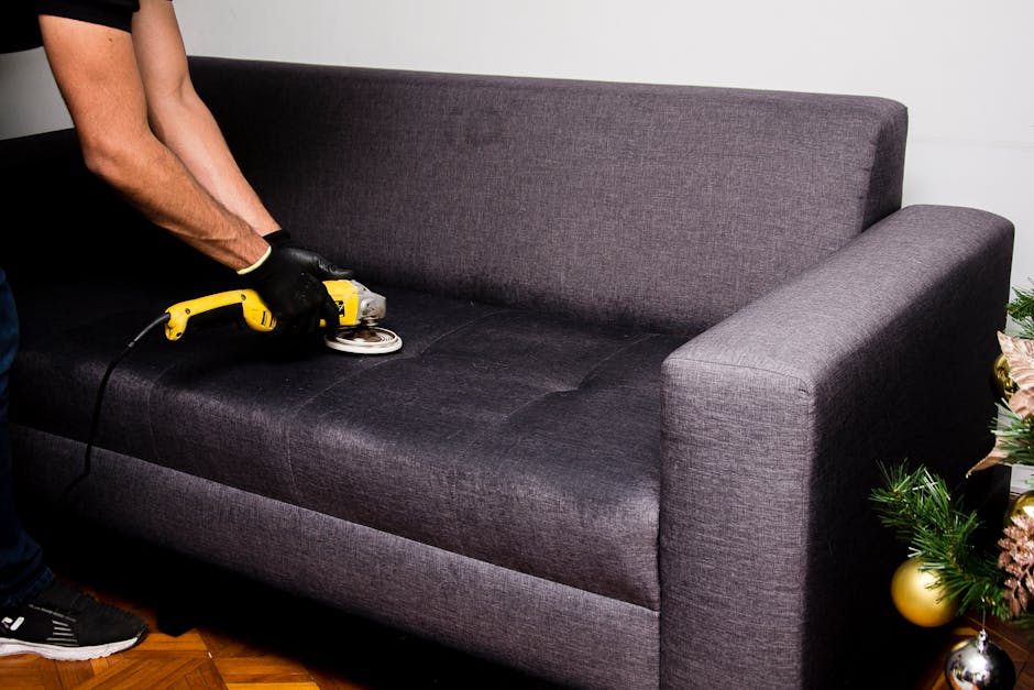 A person using a handheld buffer machine with a yellow handle on a dark grey fabric sofa in a living room, cleaning and deep cleaning the upholstery surface. The sofa features a modern design with a s