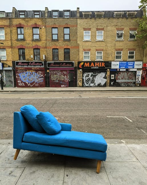 A person using a handheld buffer machine with a yellow handle on a dark grey fabric sofa in a living room, cleaning and deep cleaning the upholstery surface. The sofa features a modern design with a s